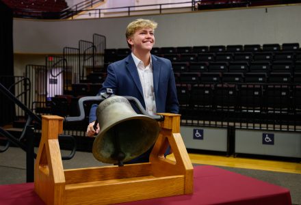 An Elon University student smiles as he stands behind a large ceremonial bell during the Opening Day ceremony.
