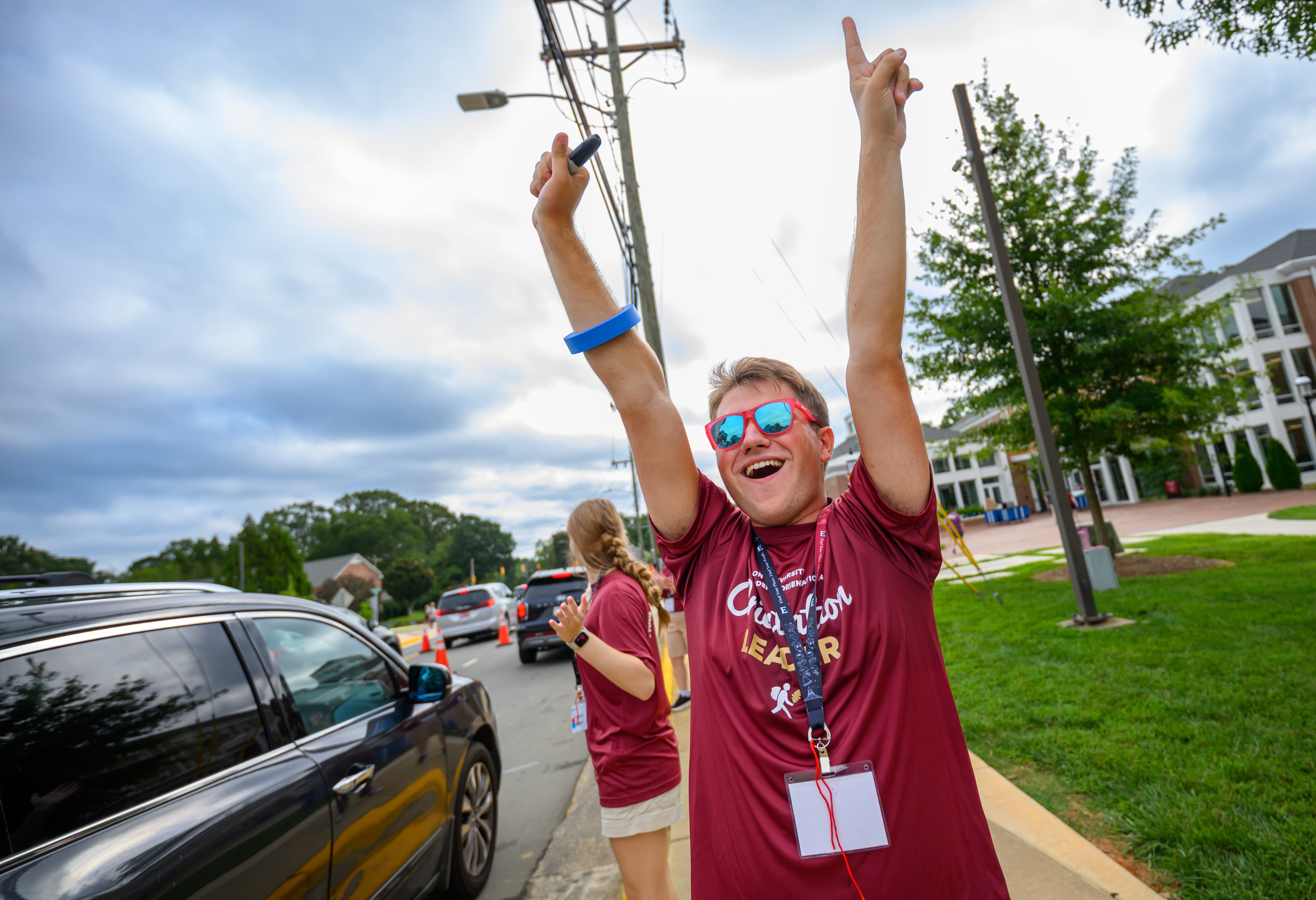 An orientation leader wearing red sunglasses raises both arms while welcoming arriving students and families.
