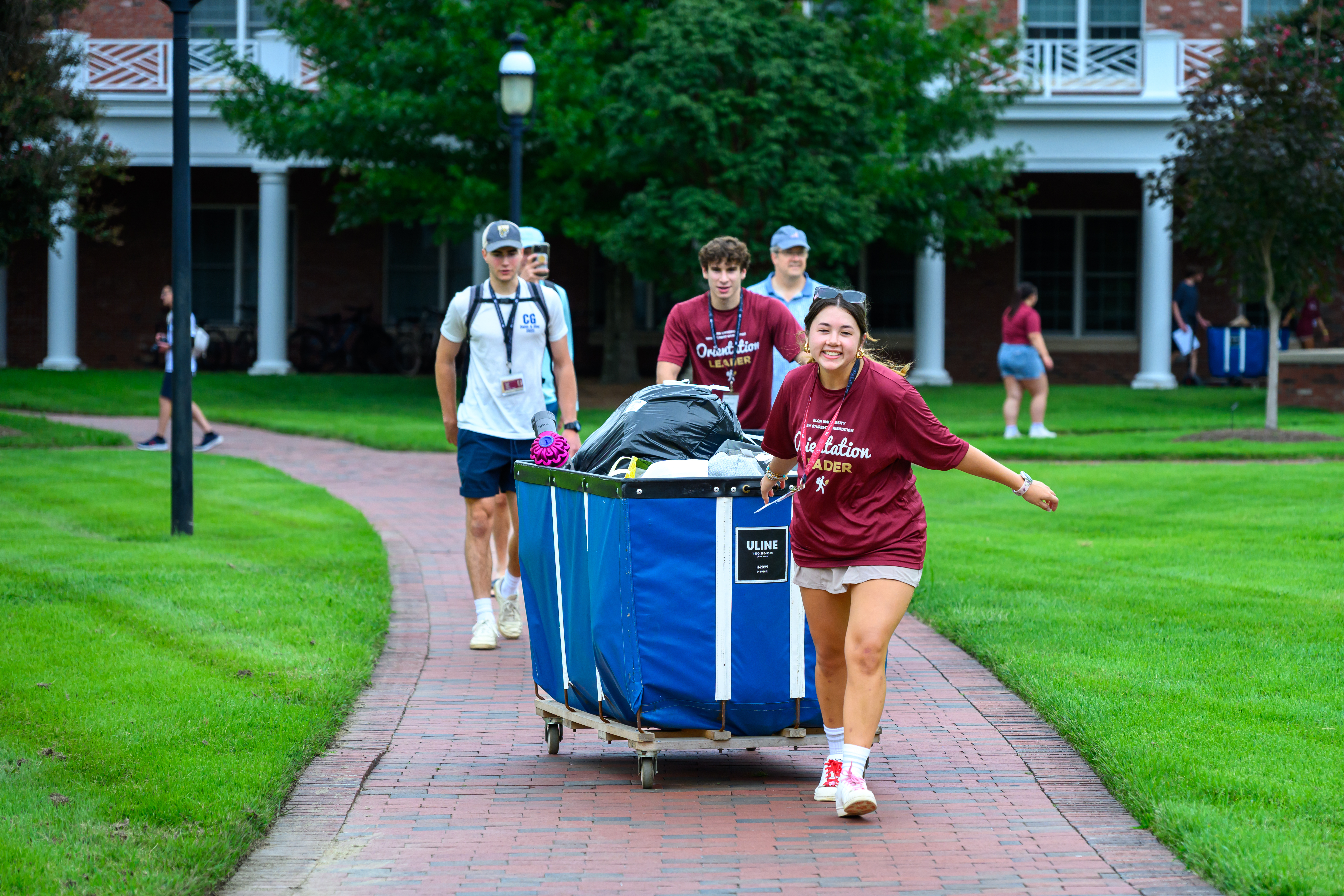 Orientation leaders push a large blue cart filled with a student’s belongings along a brick walkway during Move-In Day.