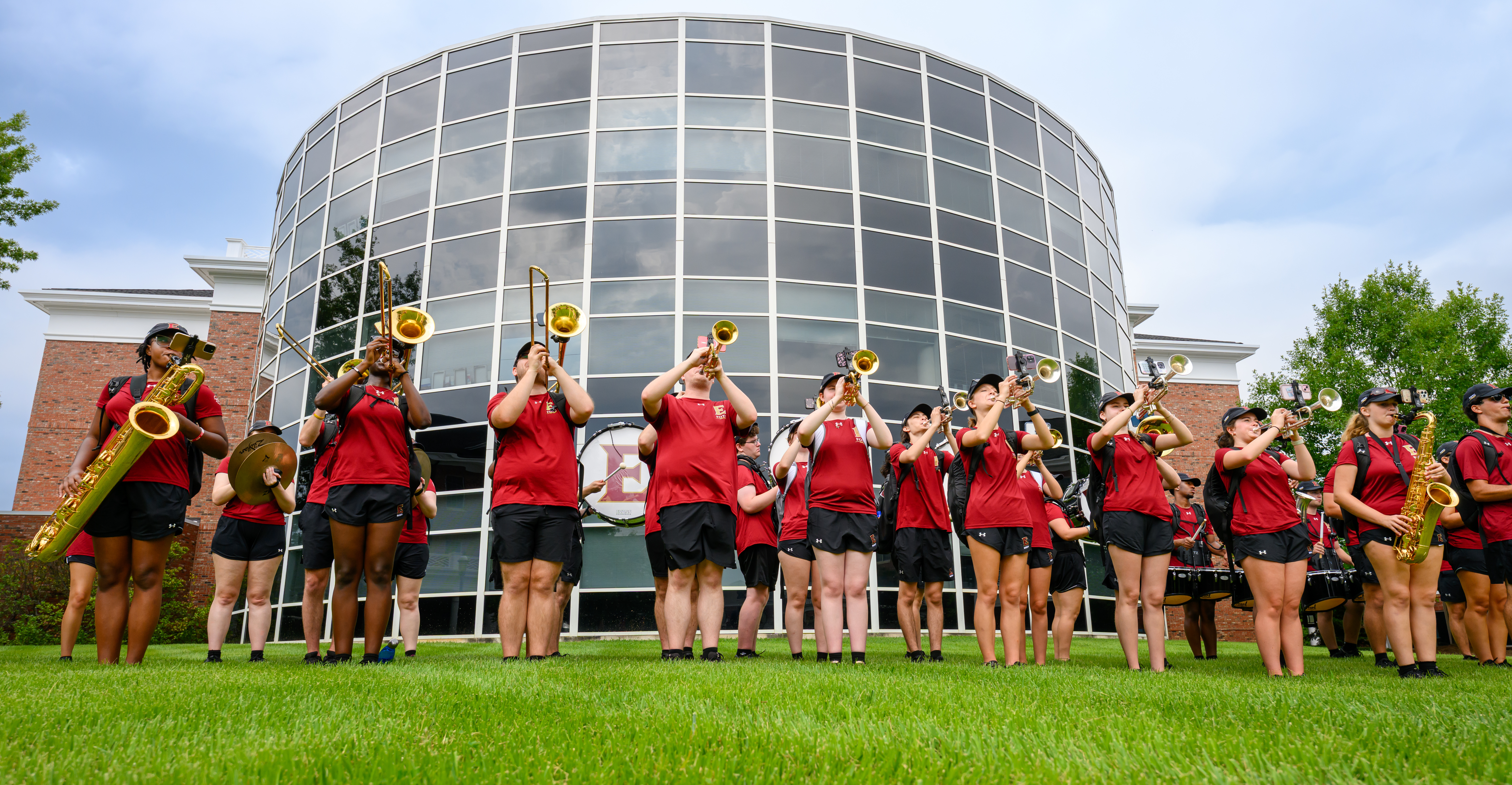 Members of the Elon marching band perform with brass and percussion instruments outside a campus building.