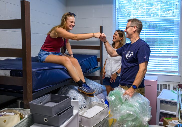 A student sitting on a residence hall bed fist-bumps a family member as they unpack belongings during Move-In Day.