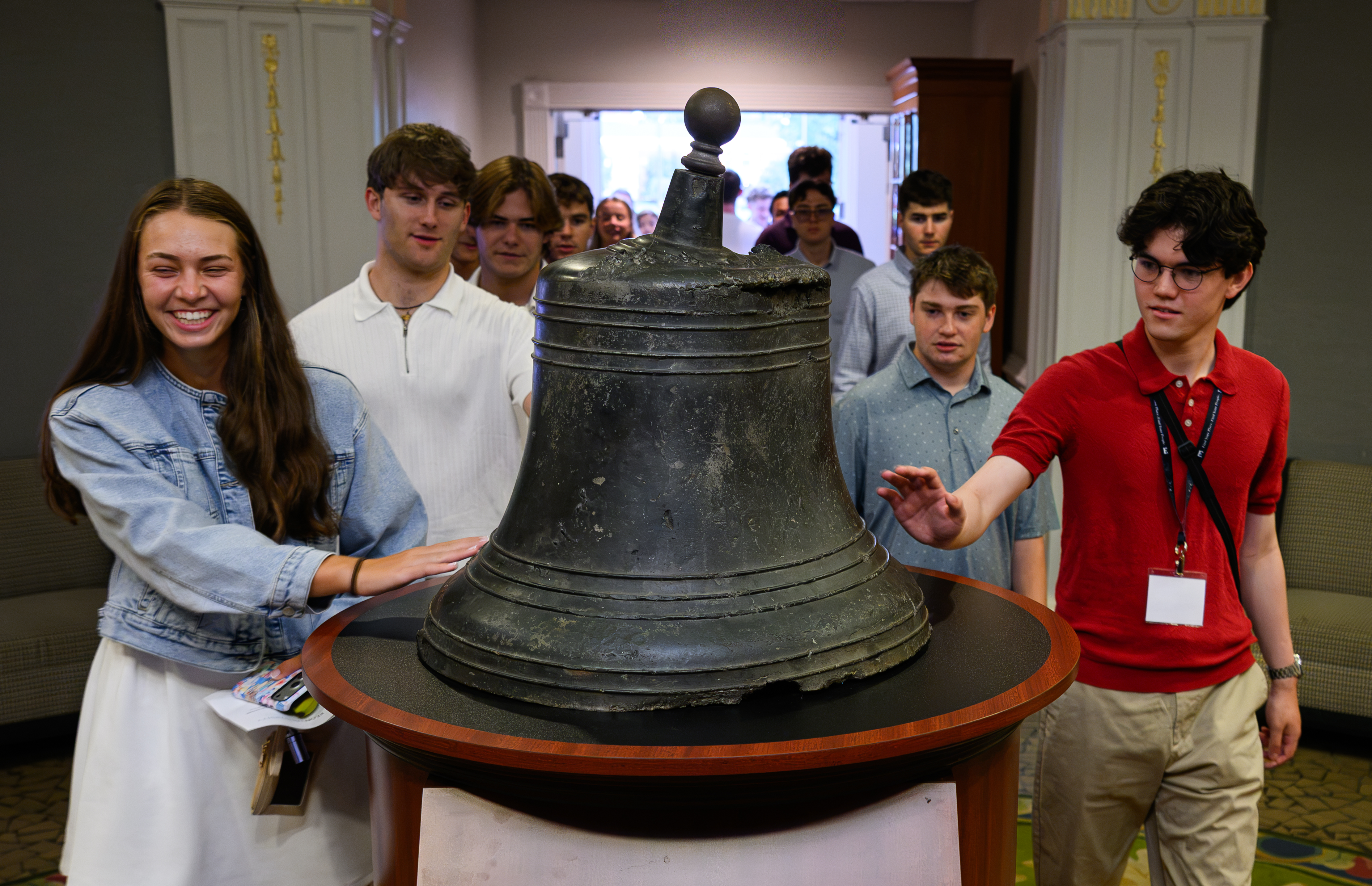 Students gather around a large historic bell, with two students reaching toward it as others watch from behind.