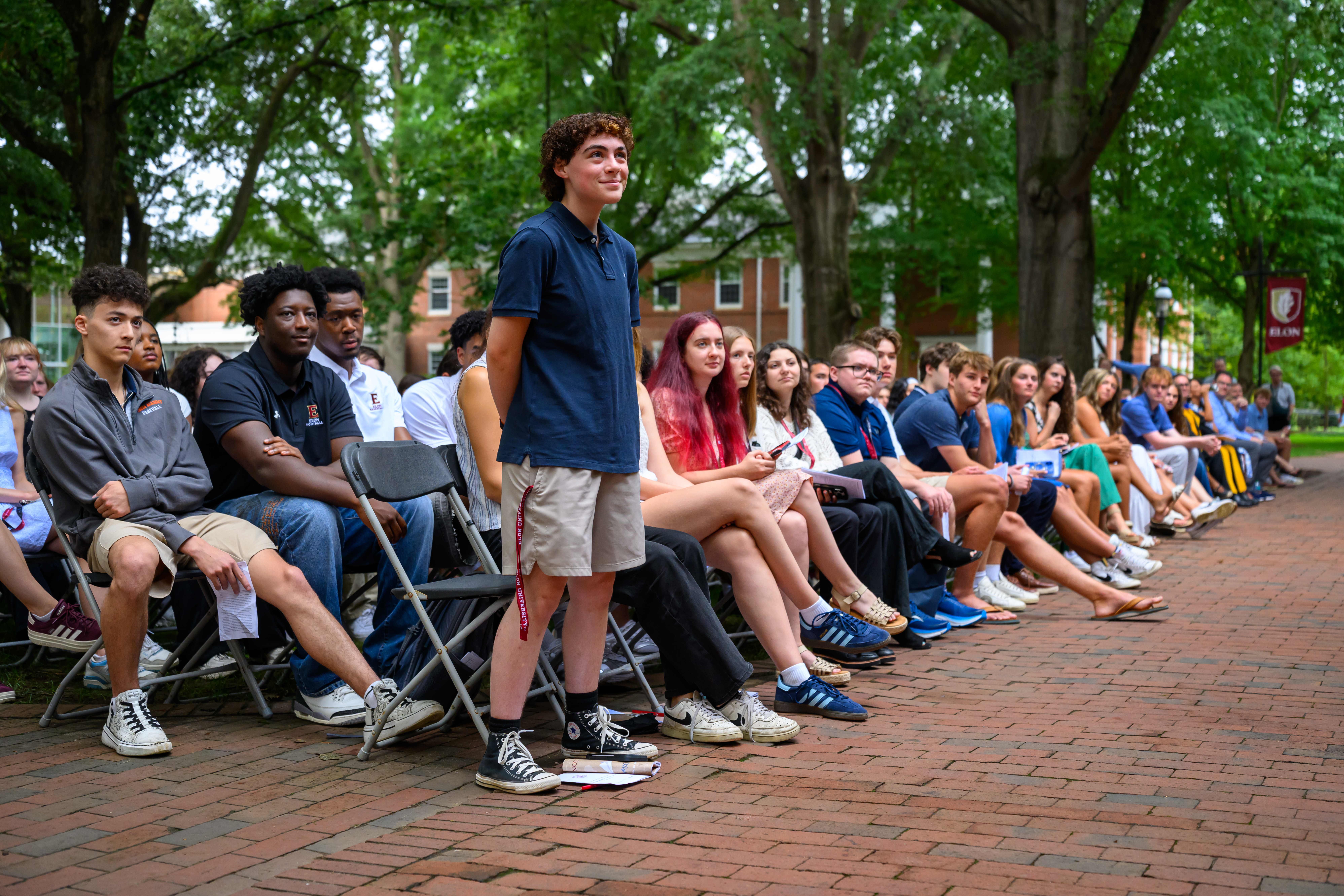 A student stands among seated classmates during New Student Convocation on a brick walkway beneath the trees.