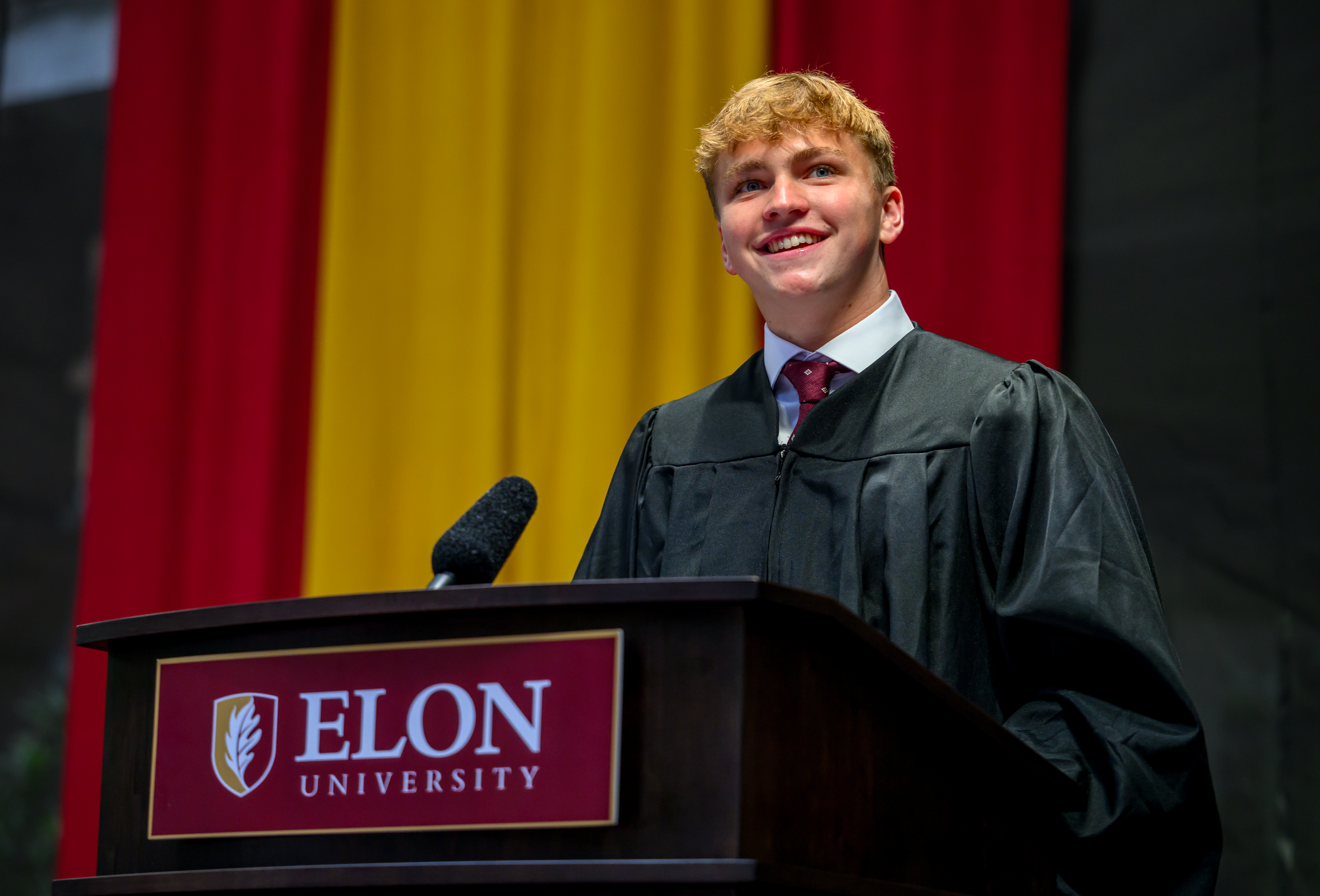 A student in academic regalia smiles while speaking from an Elon University podium against maroon and gold drapes.