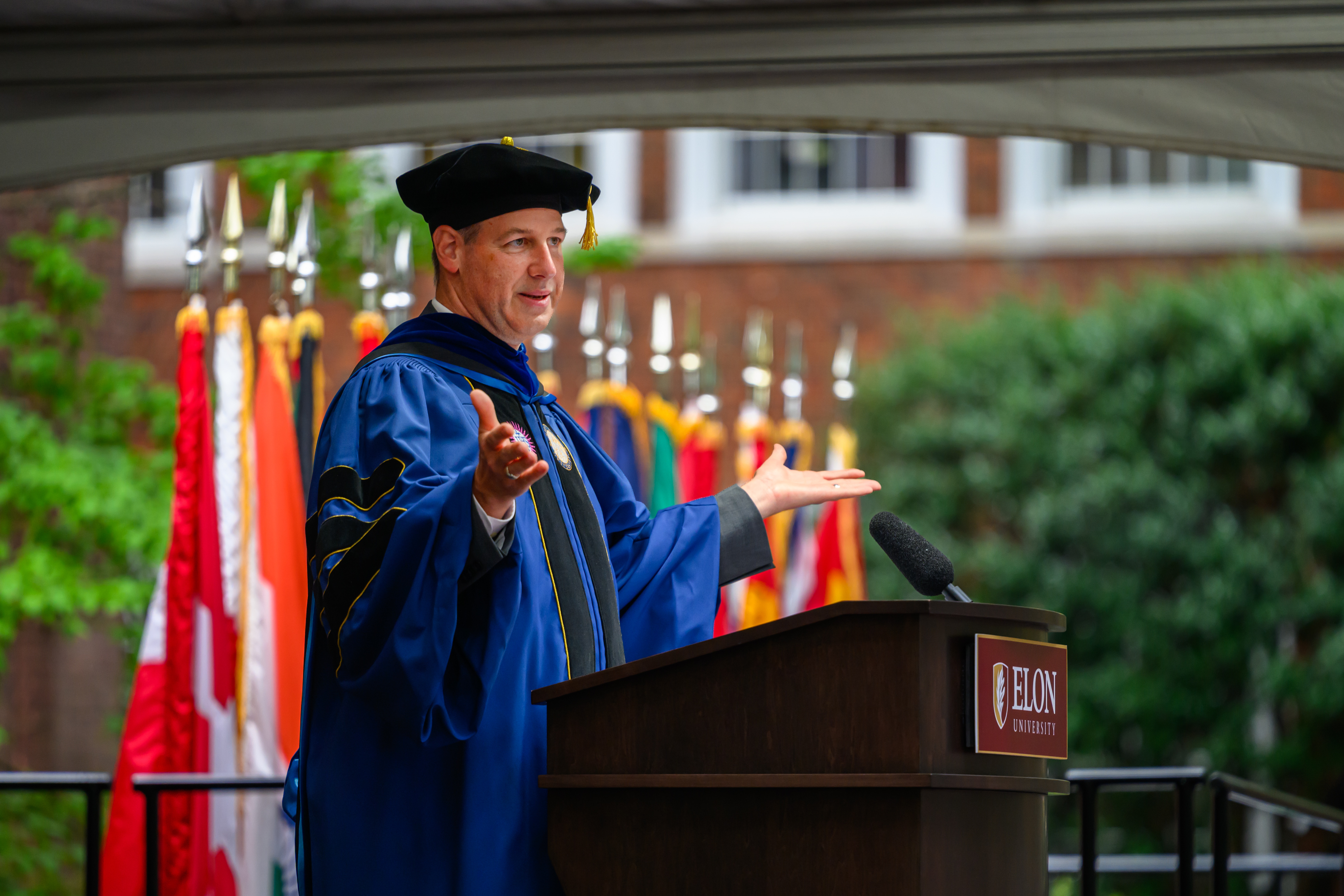 Jon Dooley in blue academic regalia gestures with both hands while speaking at an outdoor Elon University podium.