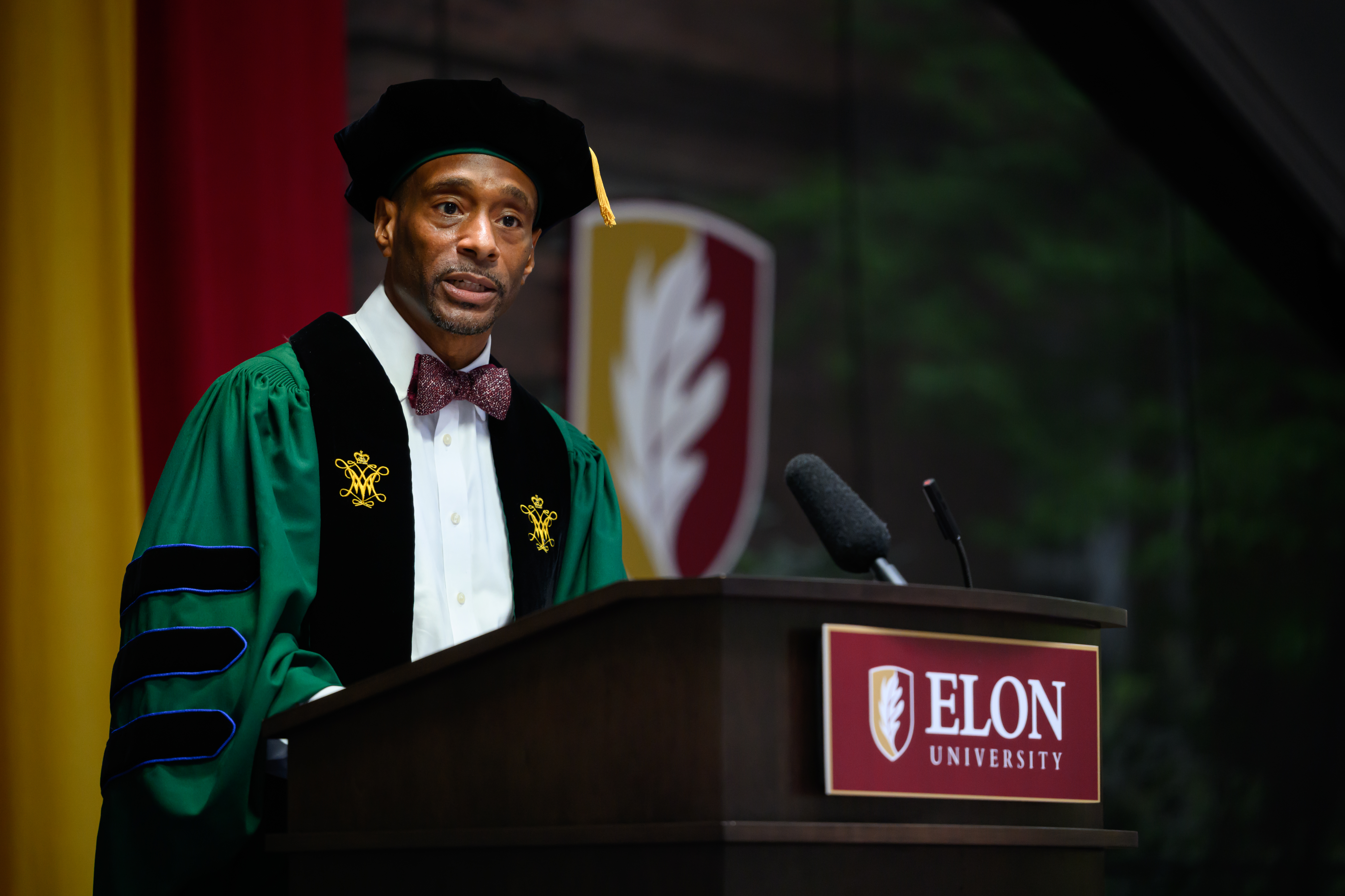 Randy Williams in green academic regalia and a bow tie speaks from an Elon University podium during New Student Convocation.