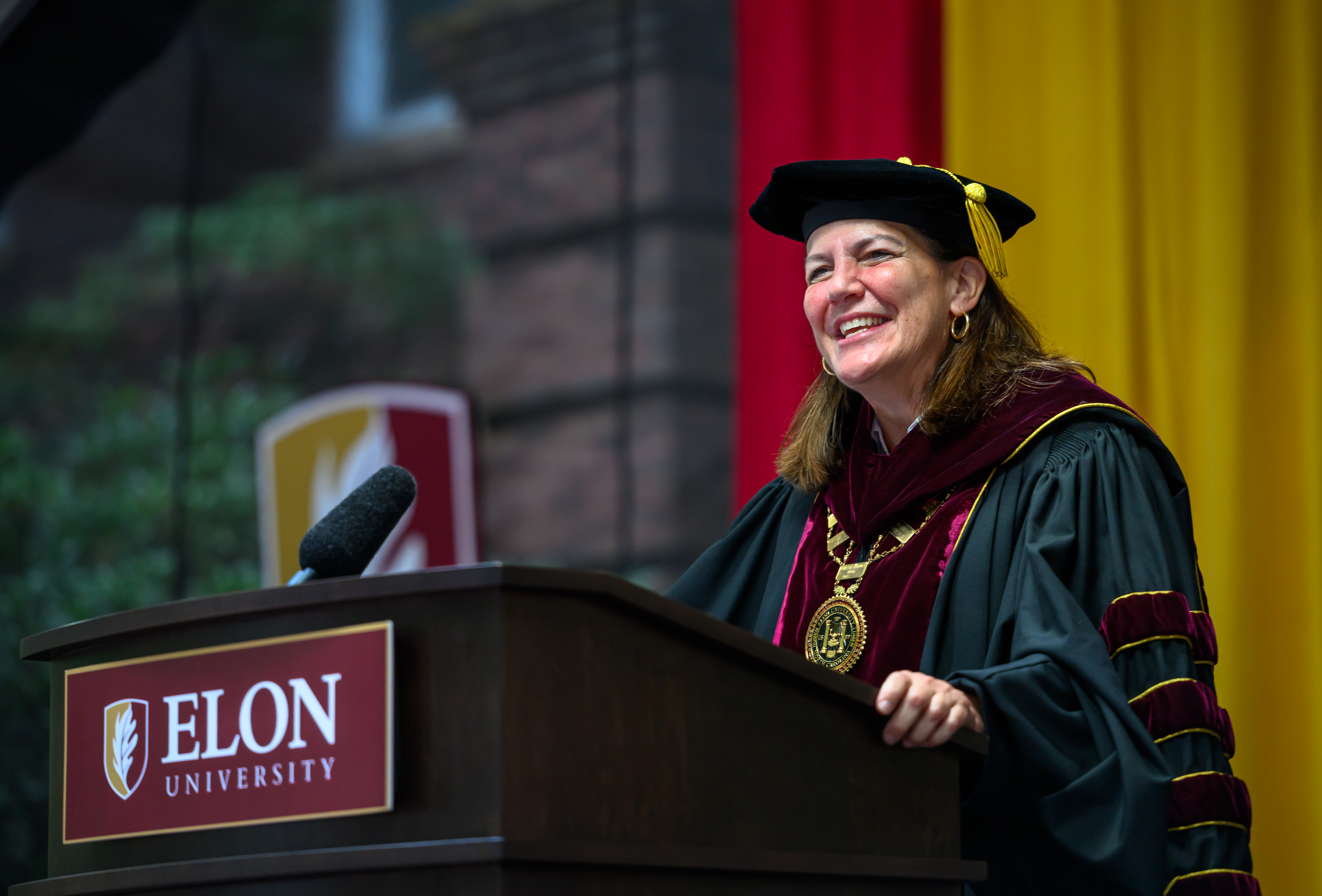 Elon University President Connie Ledoux Book smiles while speaking at a podium in academic regalia during New Student Convocation.