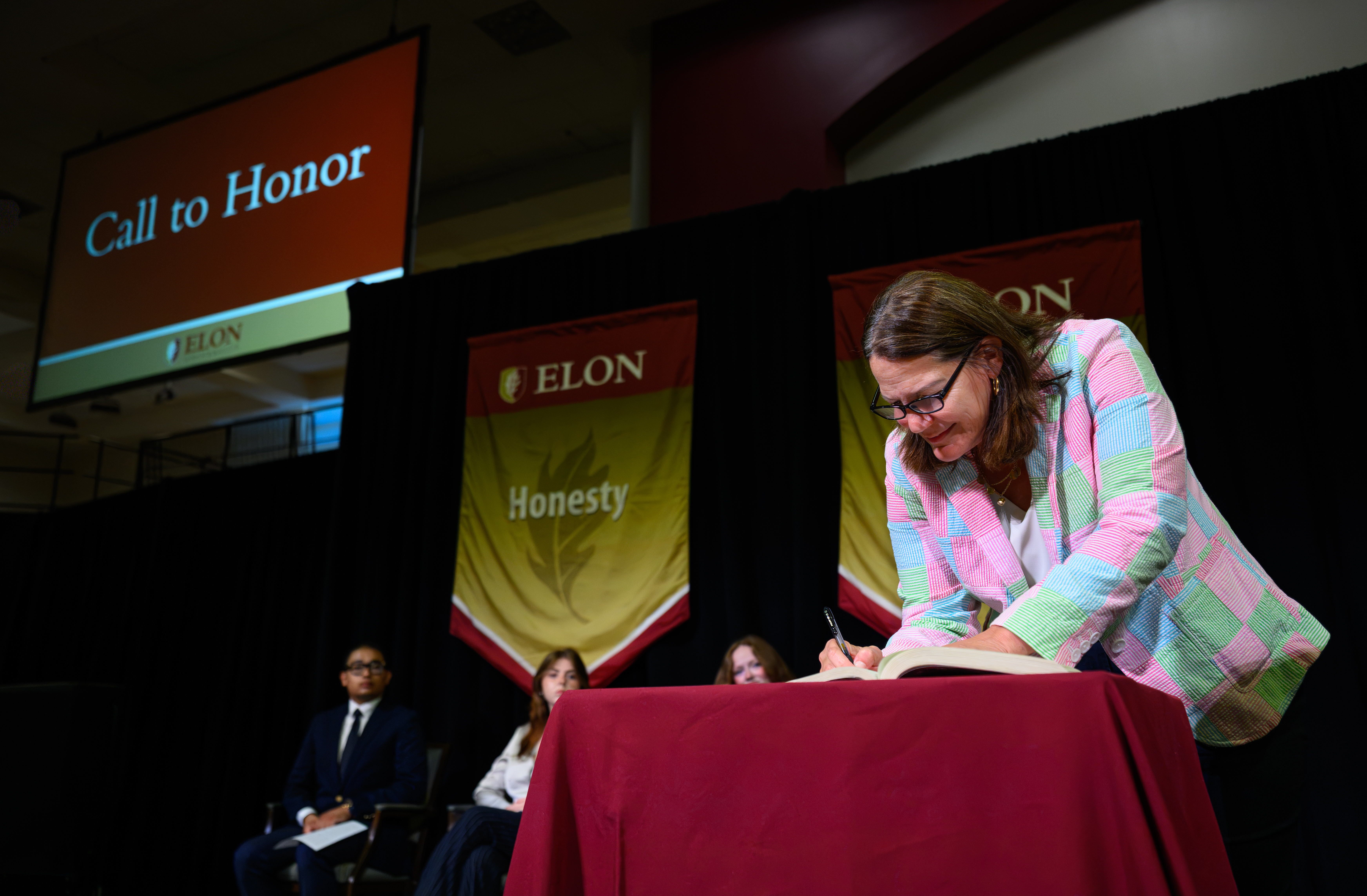 A woman signs ������Ƶ’s honor book during the Call to Honor ceremony, with an “Honesty” banner and event screen behind her.