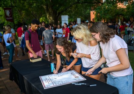 Students sign an ������Ƶ honor pledge poster at an outdoor table as a crowd gathers on a brick walkway behind them.