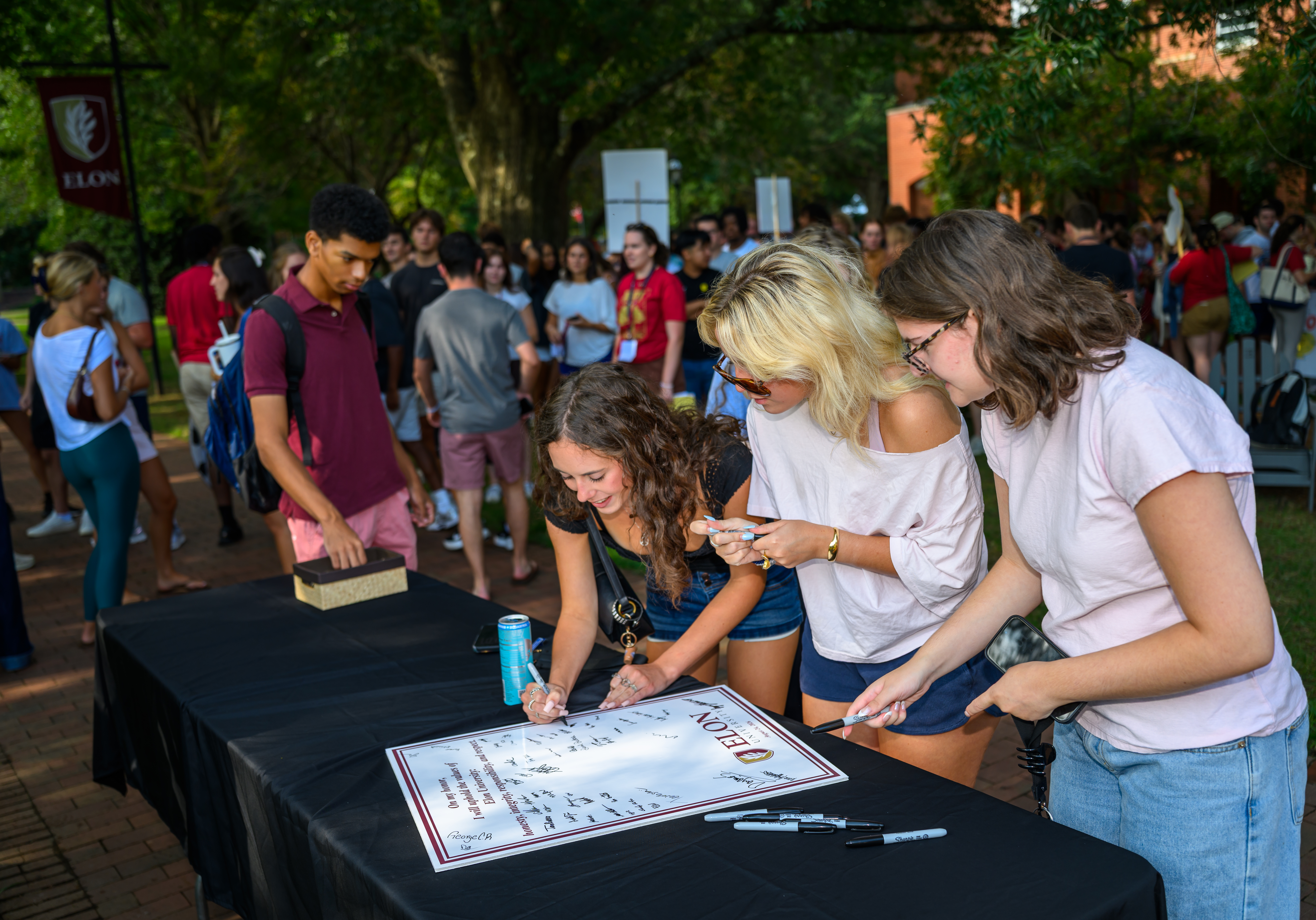 Students sign an Elon honor pledge poster at an outdoor table as a crowd gathers on a brick walkway behind them.