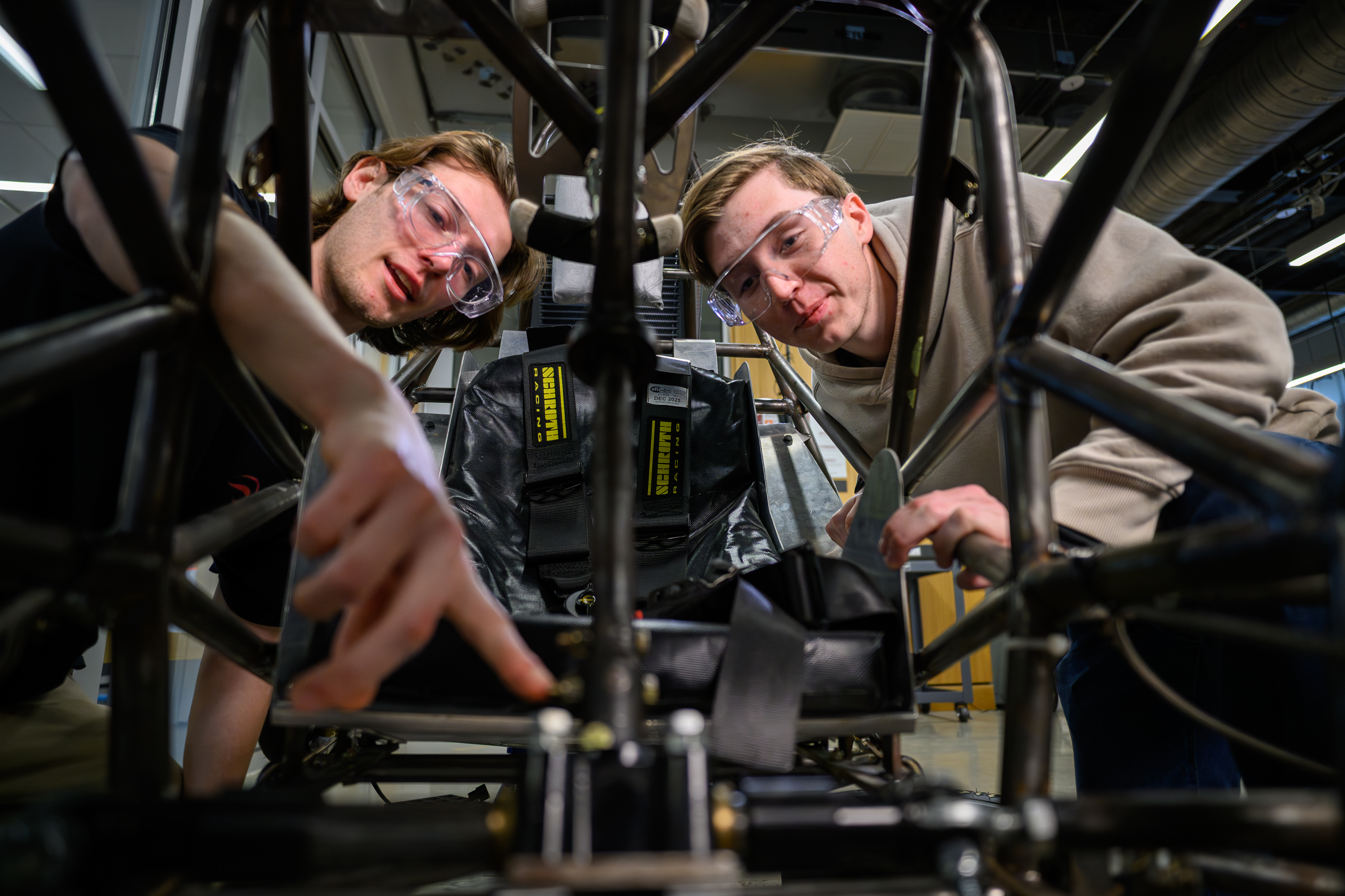Two students wearing safety glasses work together on mechanical components inside a vehicle or engineering project.