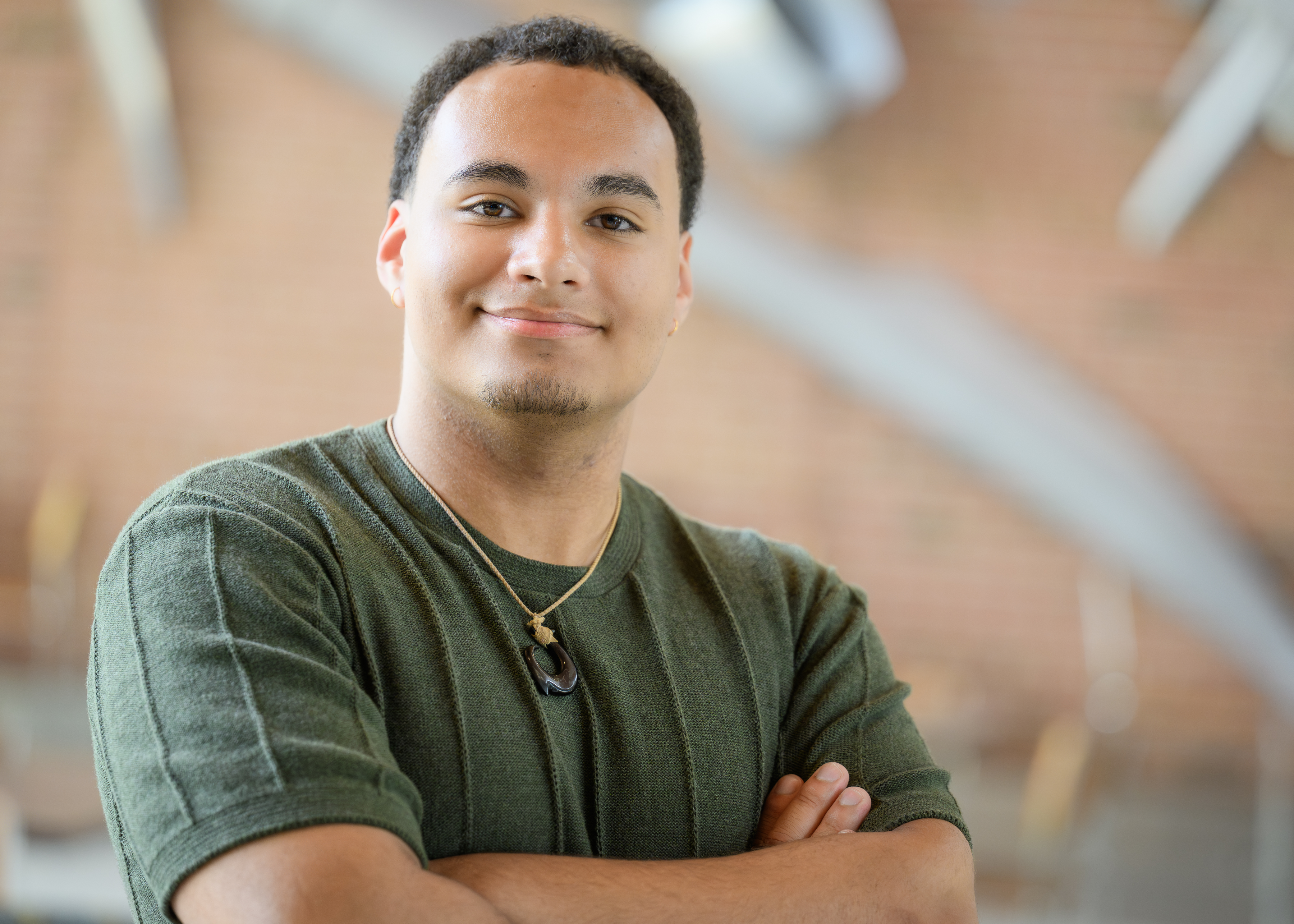 Young man in a green shirt and necklace stands with his arms crossed, smiling slightly against a softly blurred indoor background.