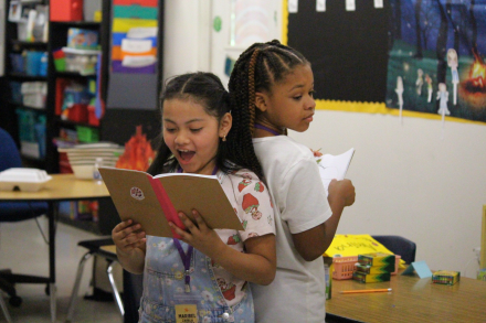 Two students stand back-to-back in a classroom while reading from books.