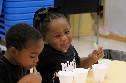 Two students sit at a classroom table with cups and straws as one smiles toward the camera.