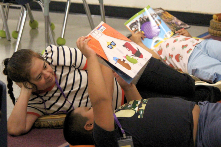 Students and a teacher lie on the classroom floor while reading colorful books together.