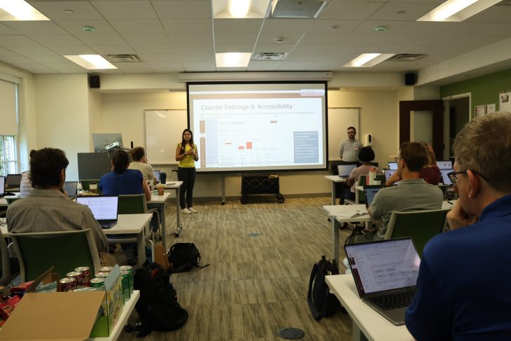 A presenter discusses course settings and accessibility at the front of a classroom as attendees follow along on laptops.