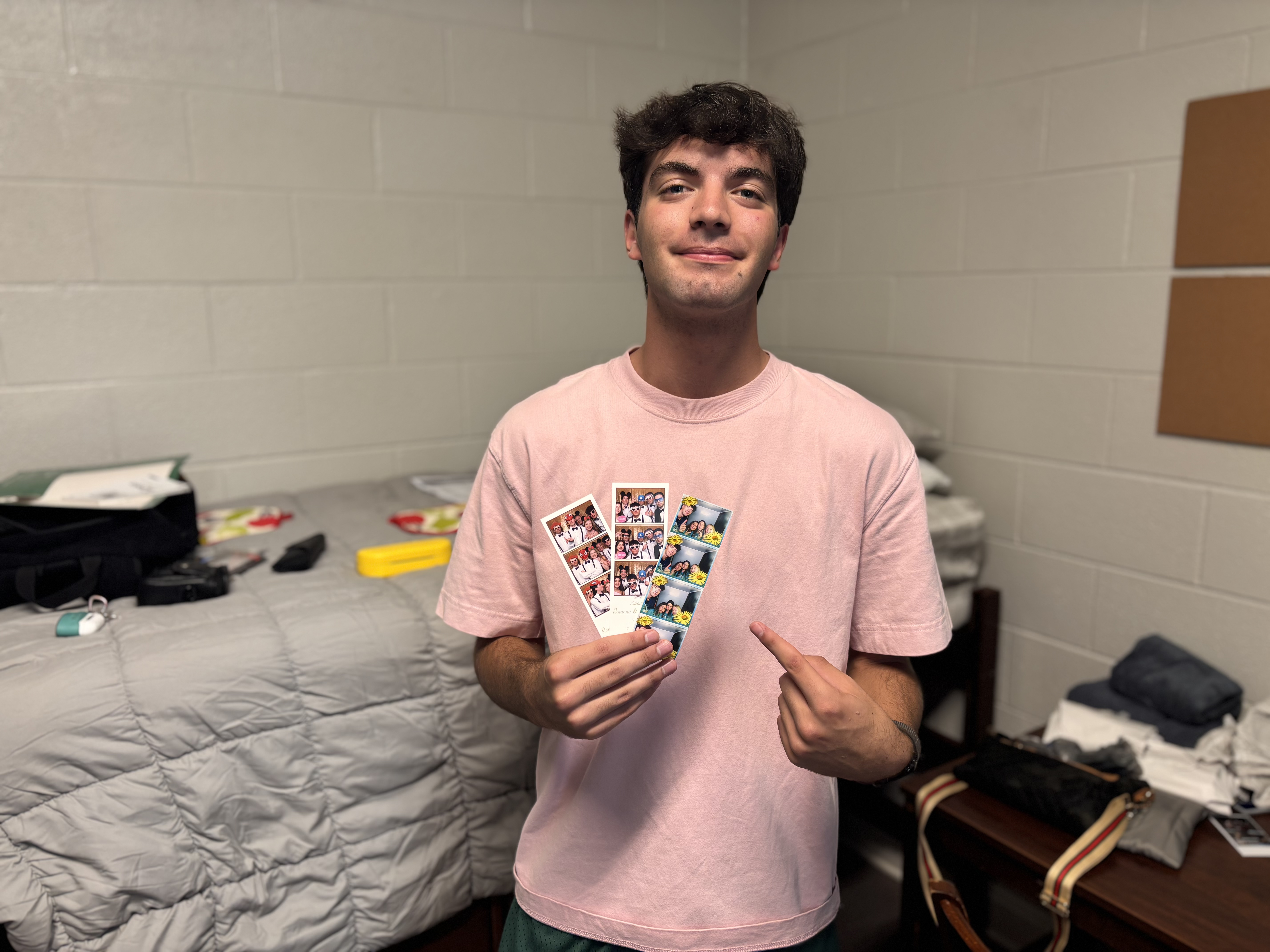 A young man holds two polaroid stips in front of a dorm room bed