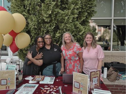 Four women smile behind an information table displaying signs, handouts and other materials at an outdoor event.