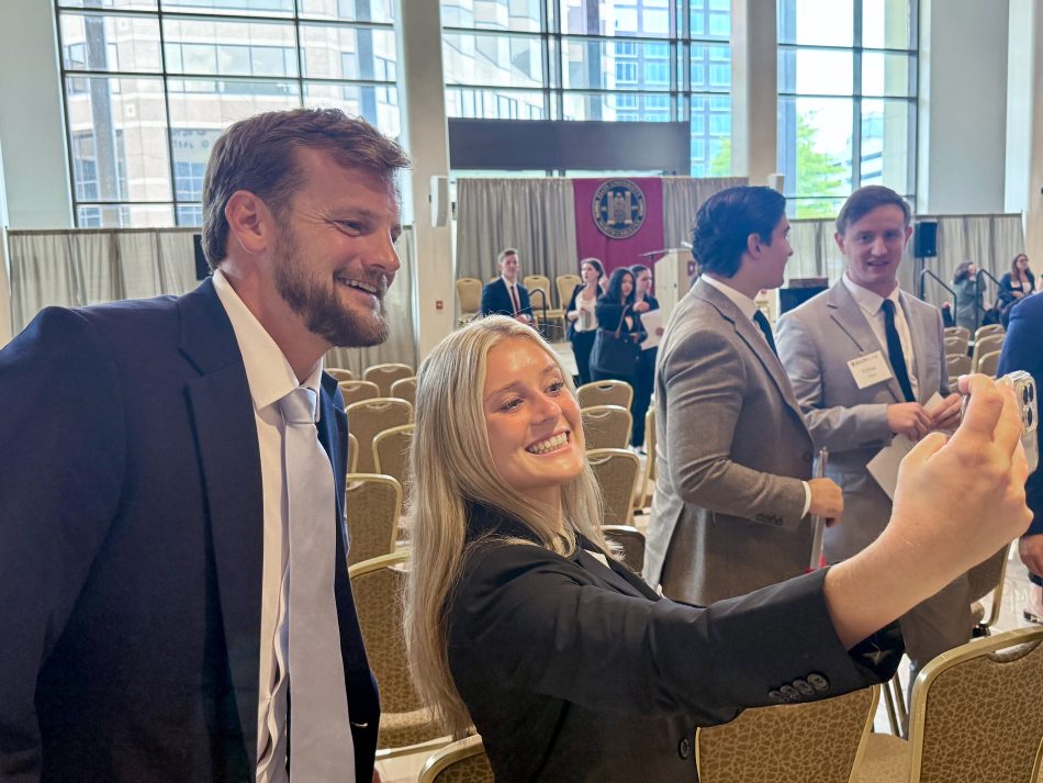 Two students taking a selfie, smiling. The large room behind them is filled with chairs and people mingling.