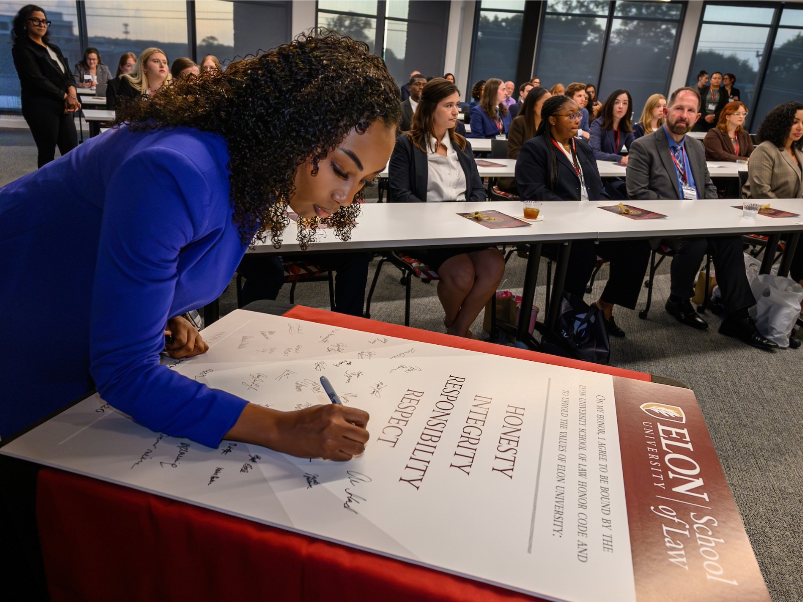 A person signing the Elon Law Honor Code. Classmates are seated in a large classroom behind her.