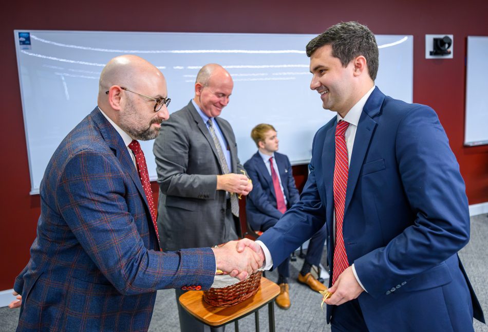 Two people shaking hands. A person behind them holds an acorn in a small gold bag.