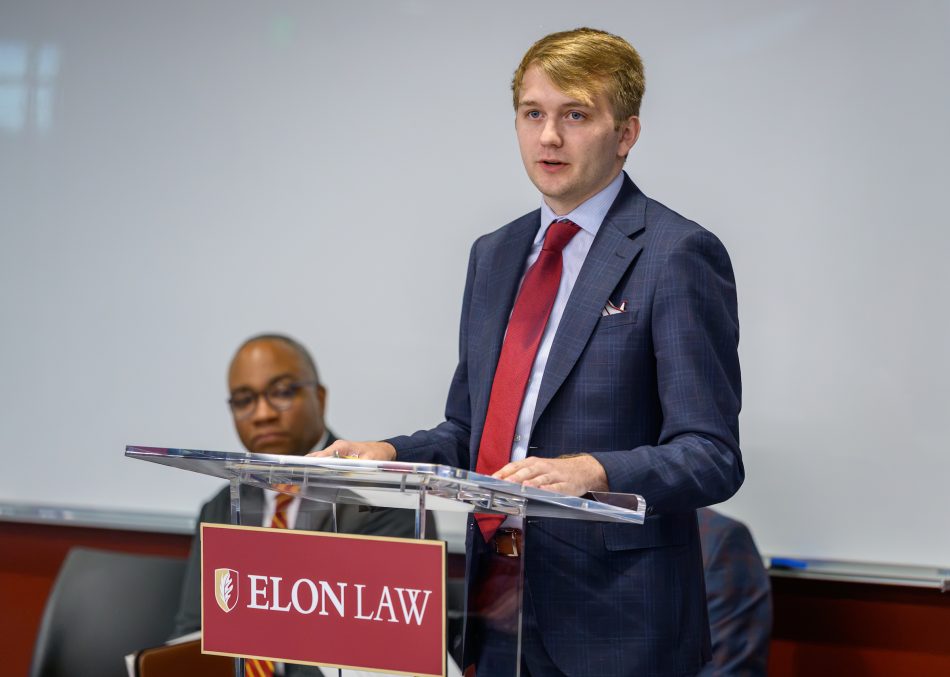 A person speaks at a podium with an Elon Law plaquard on it.