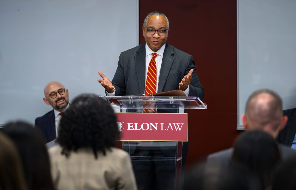 A man gestures while speaking at a podium with an Elon Law plaquard on it.