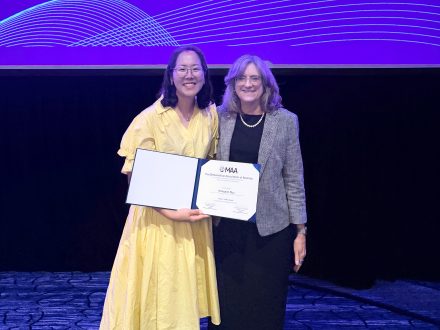 Hwayeon Ryu in a yellow dress holding her MAA award and standing with Jenna P. Carpenter in a black outfit with a grey blazer at the MAA award.