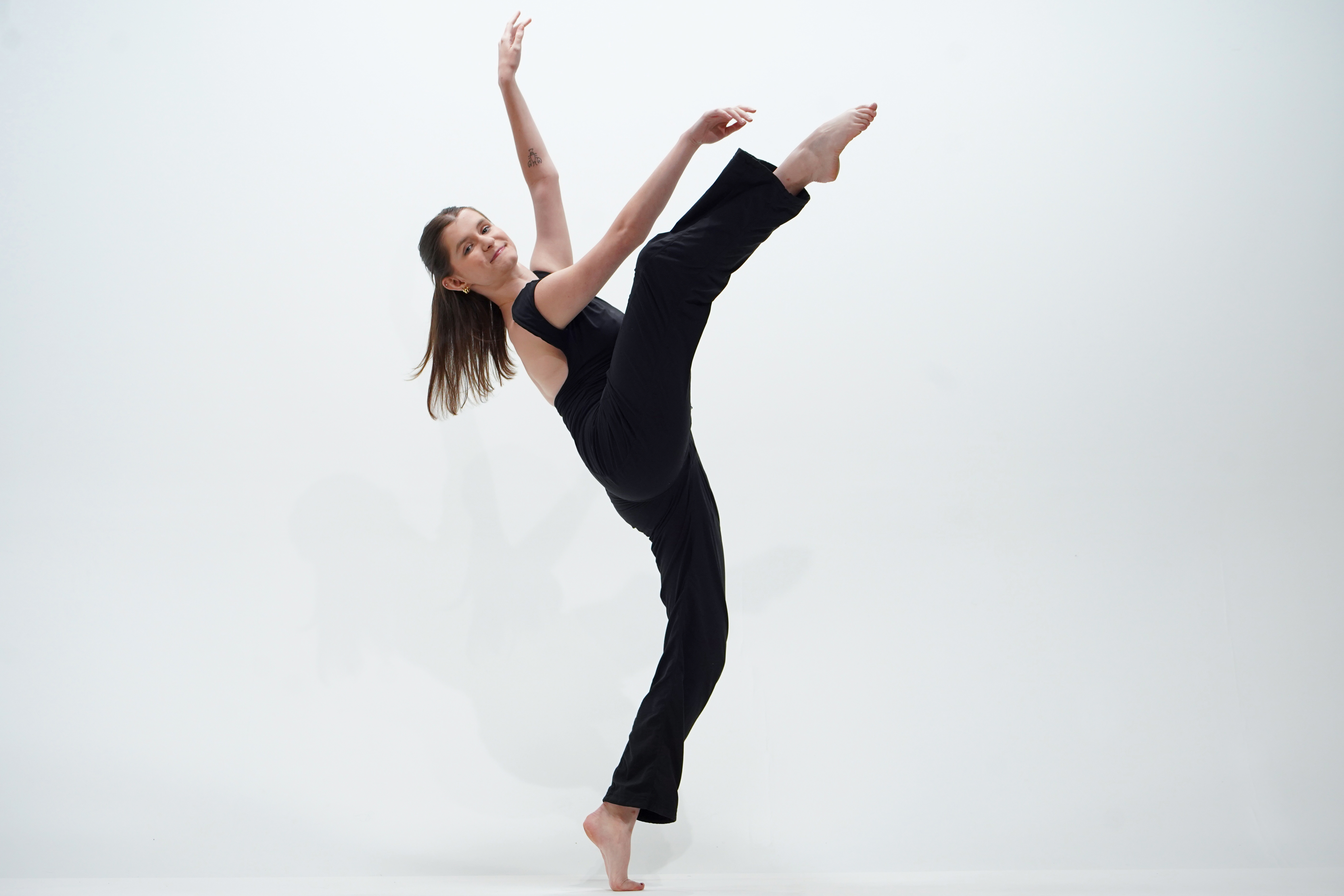 Barefoot dancer dressed in black balances on one leg while extending her other leg high against a white studio background.