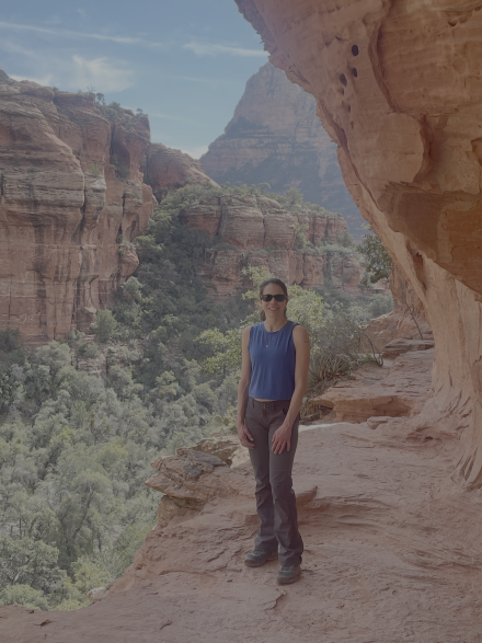 Kaitlin Henze ’12 stands beneath a red-rock overhang during a hike, with towering sandstone cliffs and a forested canyon behind her.
