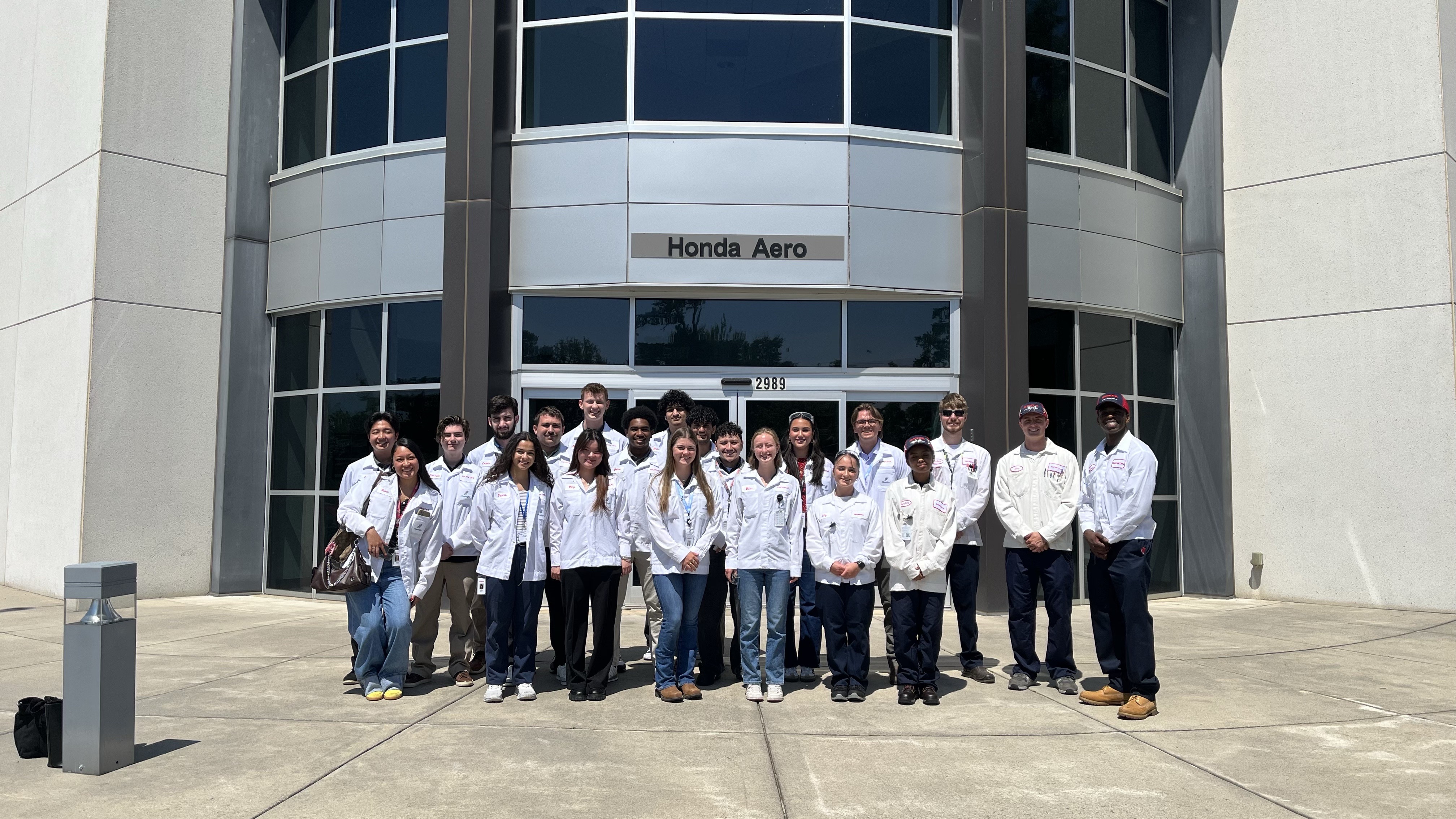 A group of Honda Aircraft interns wearing white work coats stand together outside the entrance to the Honda Aero facility.