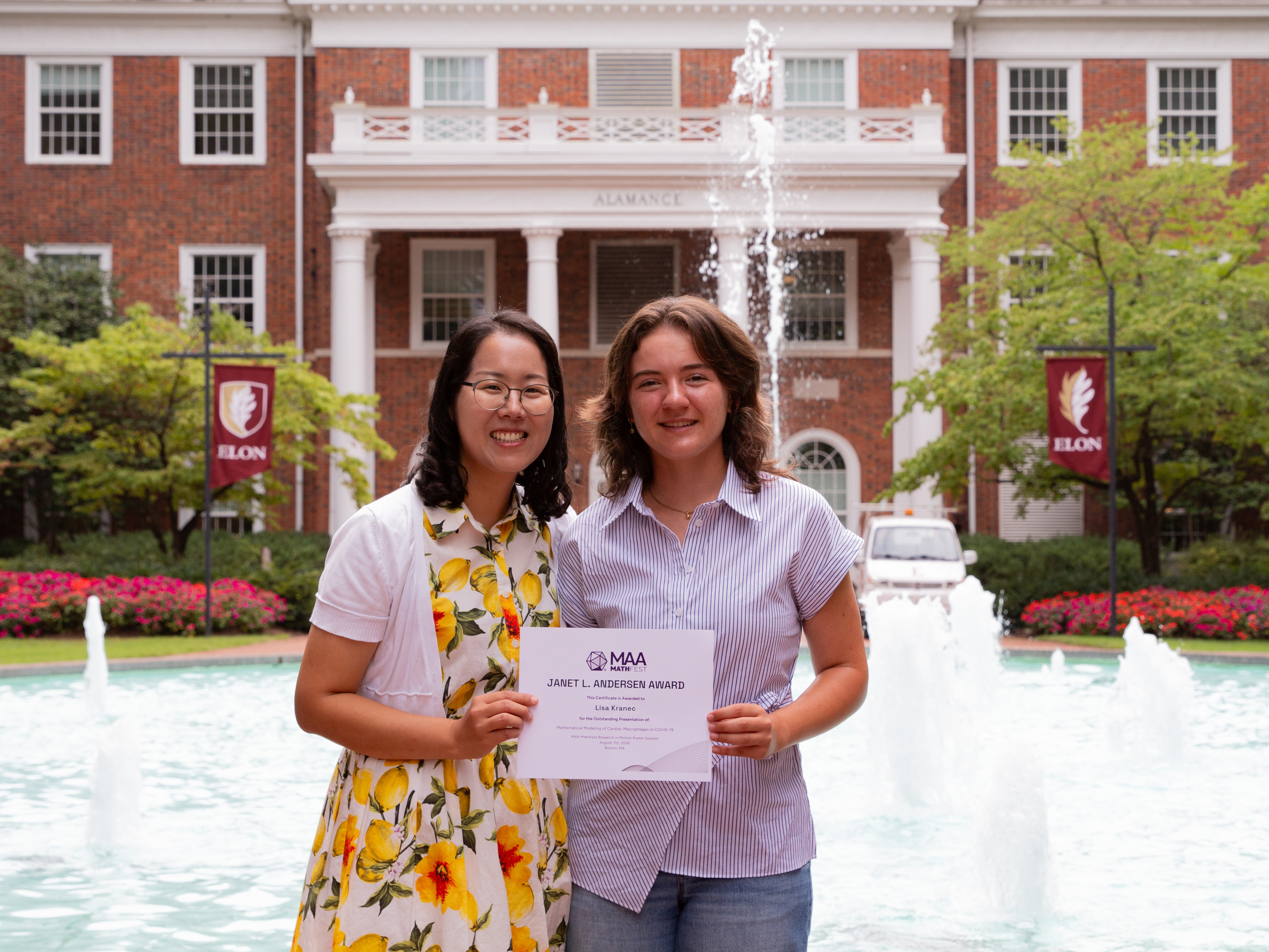 Student Lisa Kranec in a white shirt standing with associate professor Hwayeon Ryu who is wearing a dress with lemons on it posing in front of the fountain at Alamance building holding Lisa's award certificate.