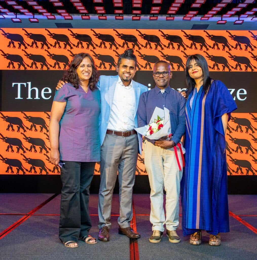 Four people pose together onstage at an awards ceremony, with one holding a bouquet of flowers.