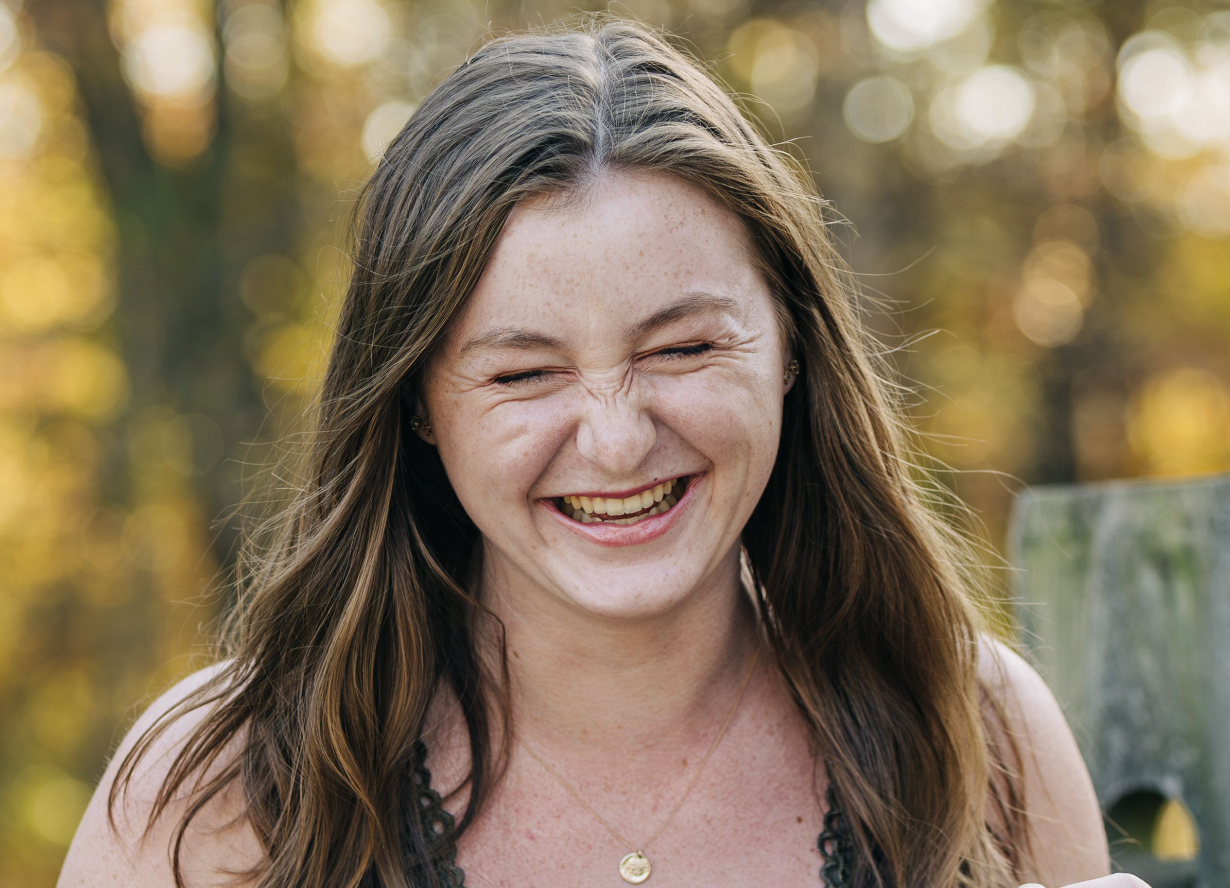Young woman laughs with her eyes closed outdoors, with long brown hair and sunlit trees blurred in the background.