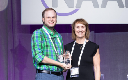 Two people stand side by side for a posed photo. The person on the left holds an award.