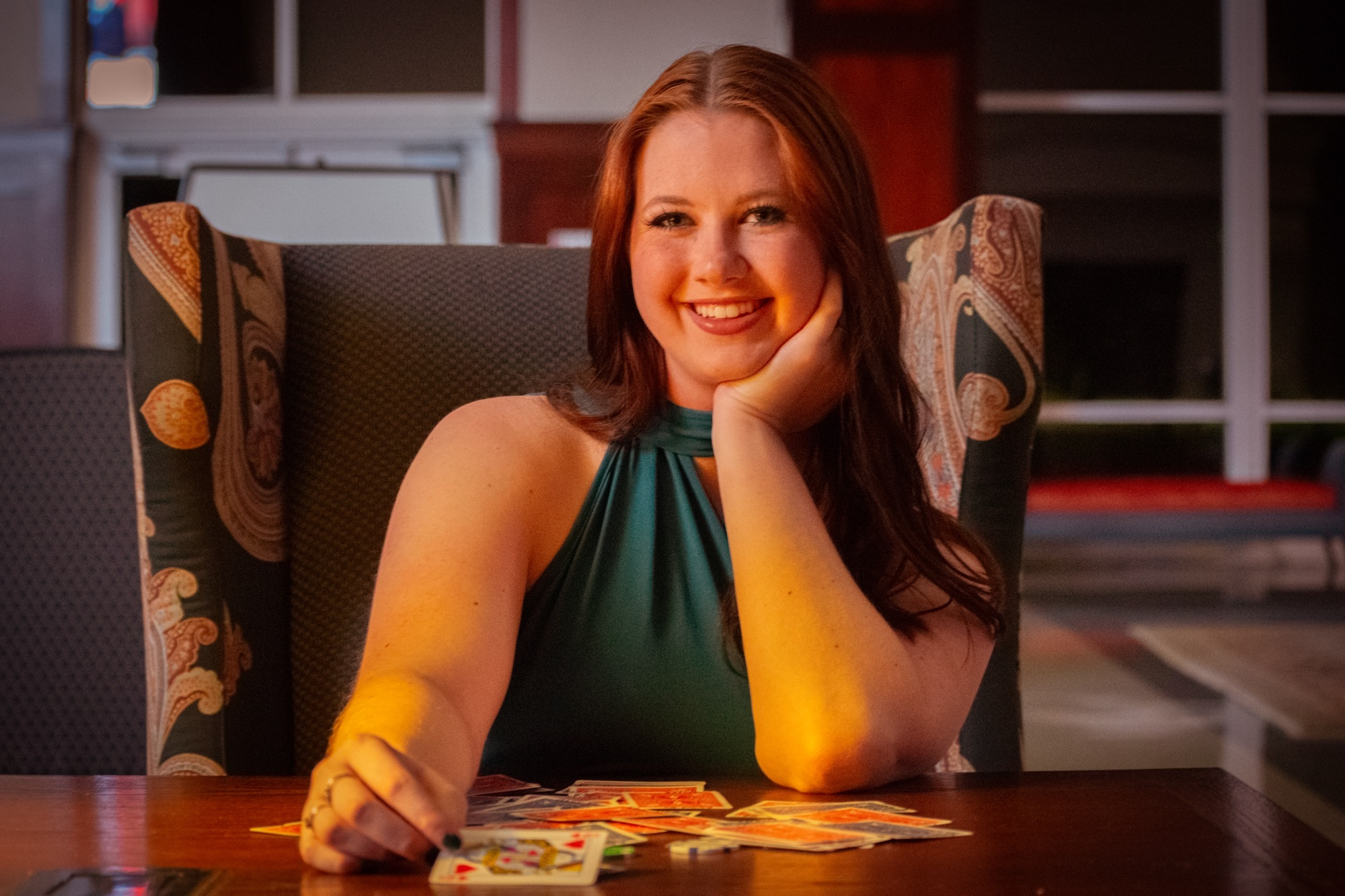 Young woman in a green sleeveless top smiles while seated at a table with playing cards spread in front of her.