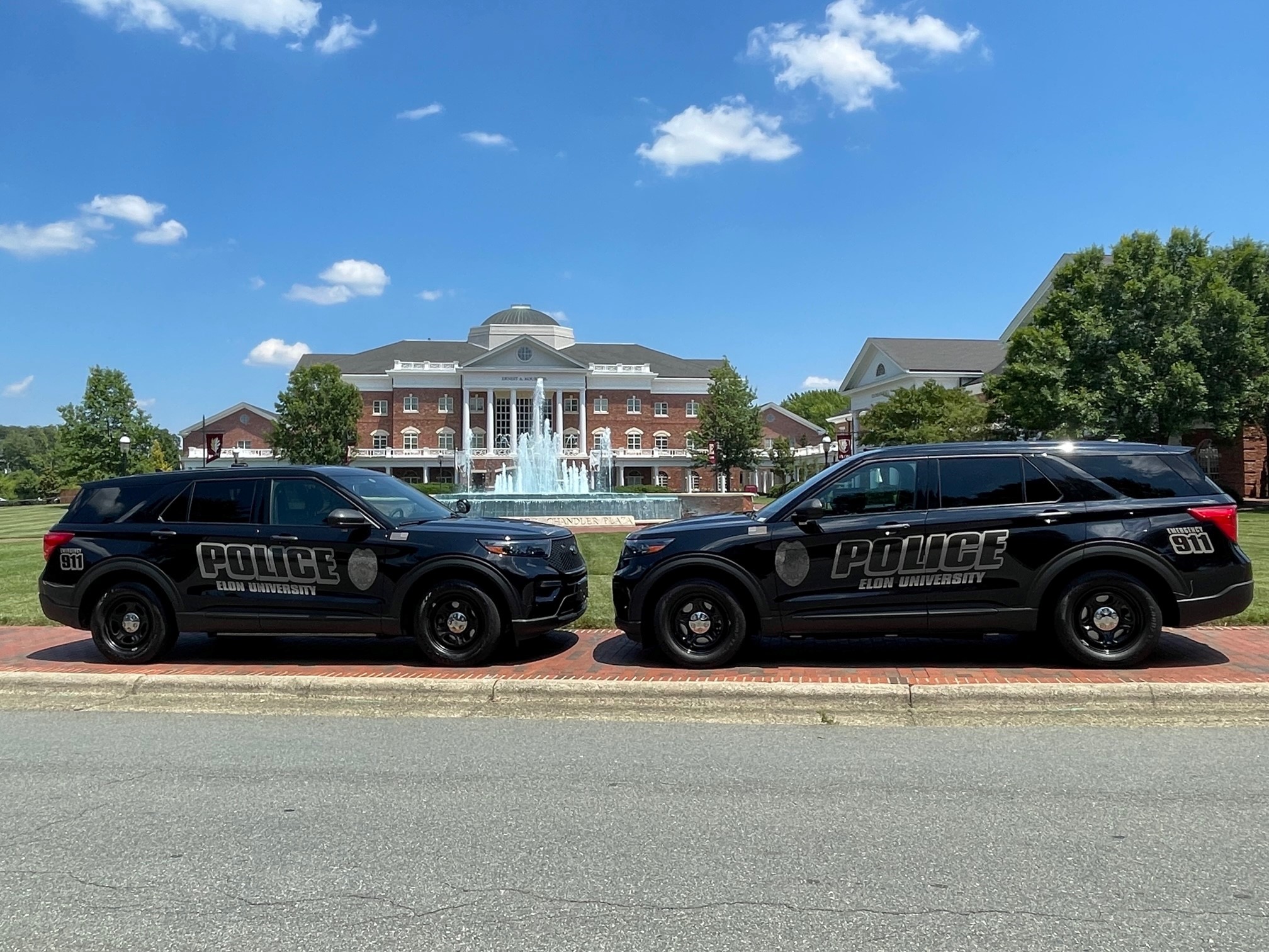 Two 51������ Police SUVs are parked nose to nose in front of Young Commons, with the fountain spraying beneath a bright blue sky.