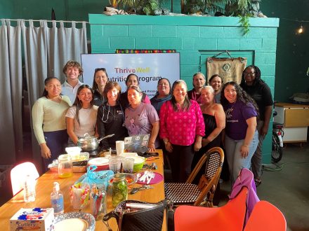 Twelve people stand together smiling for a group photo behind a long table covered with fresh vegetables, cooking ingredients, utensils and prepared food during a Thrive Well nutrition program. A banner reading “Thrive Well Nutrition Program” is displayed behind the group, and the gathering takes place in a bright community space with colorful seating and plants in the background.