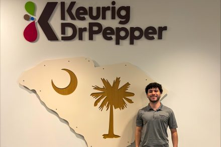 William Coughenour stands beneath a Keurig Dr Pepper sign beside a wall display shaped like South Carolina with a crescent moon and palmetto tree.