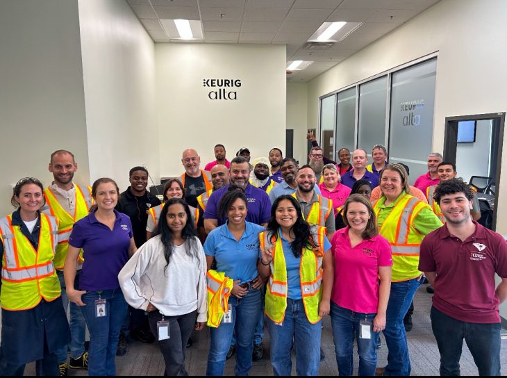 William Coughenour stands at far right with a large group of Keurig Dr Pepper employees, several wearing safety vests, inside a Keurig Alta facility.