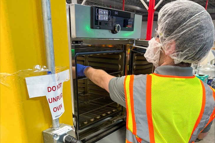William Coughenour, wearing a safety vest, hair covering and protective eyewear, works with equipment on the Keurig Dr Pepper production floor.