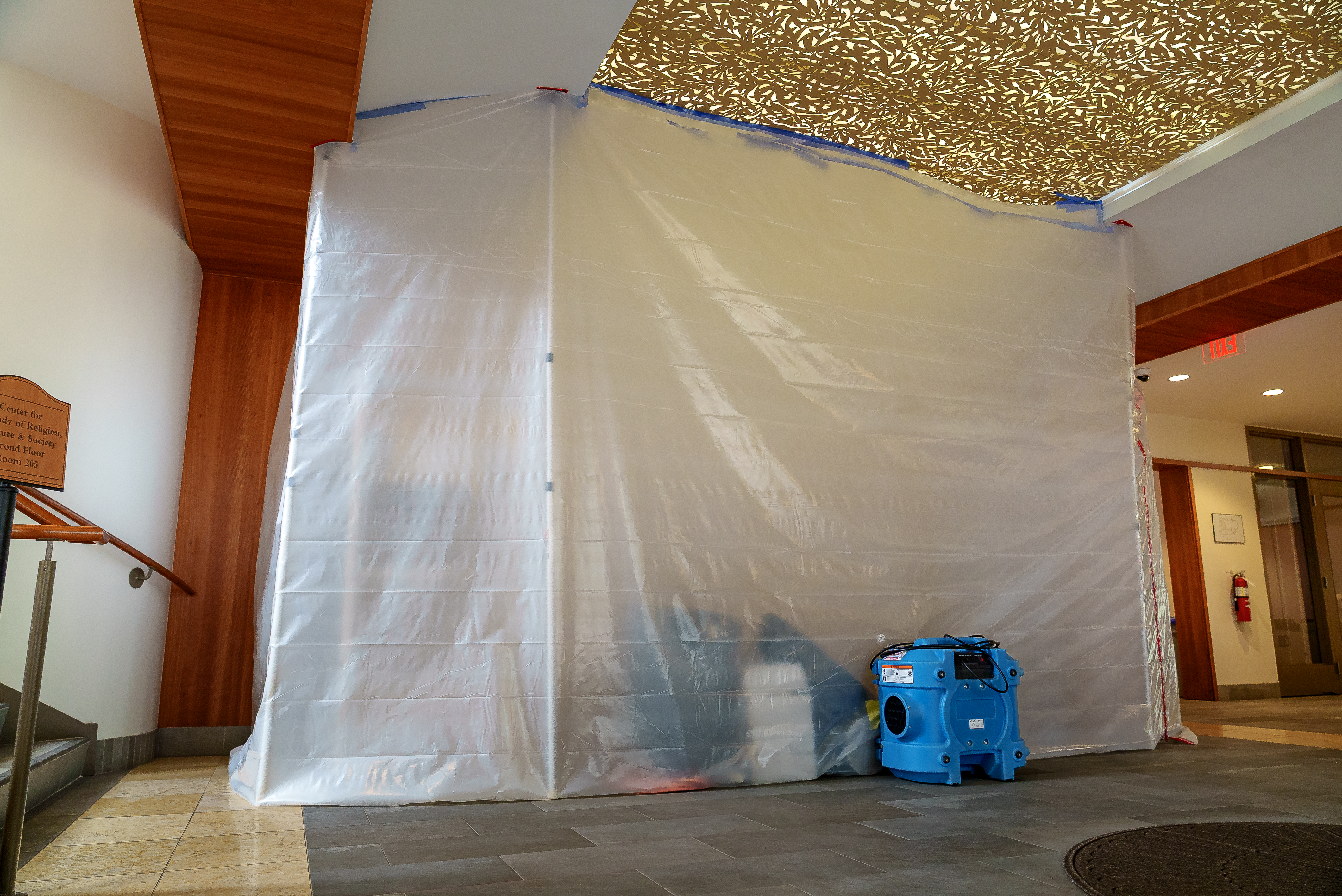Plastic containment sheeting encloses part of an interior hallway during renovation work, with a blue air filtration unit positioned outside the temporary barrier.
