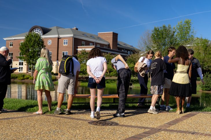 Students gather around a newly dedicated bench beside a campus pond, with two speakers and a brick building in the background.