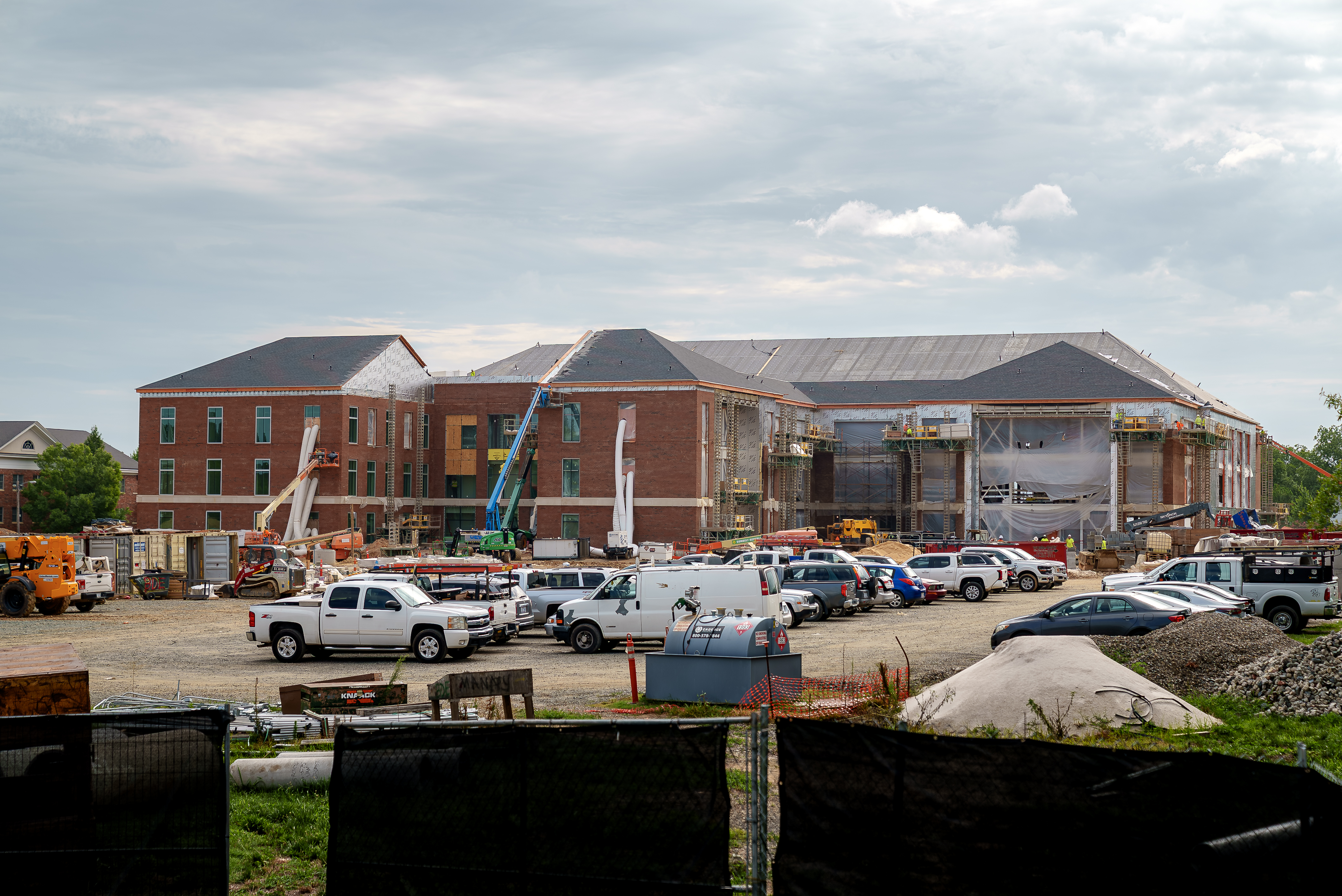 A large brick building under construction rises behind a busy worksite filled with pickup trucks, construction equipment and scaffolding. Roofing and exterior work are underway beneath a cloudy sky.