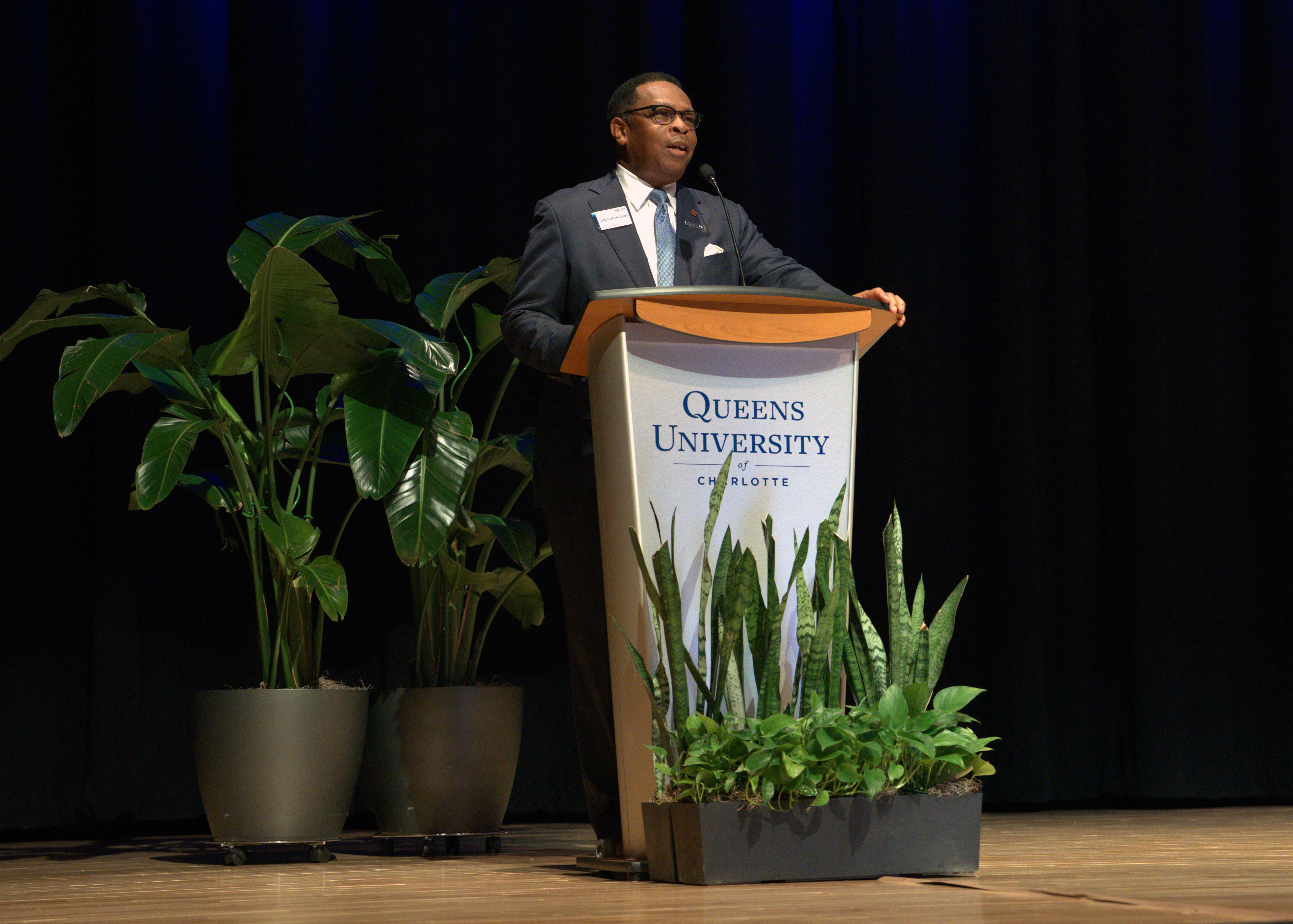 Man wearing glasses stands at podium that says Queens University of Charlotte with plants around podium on a stage.