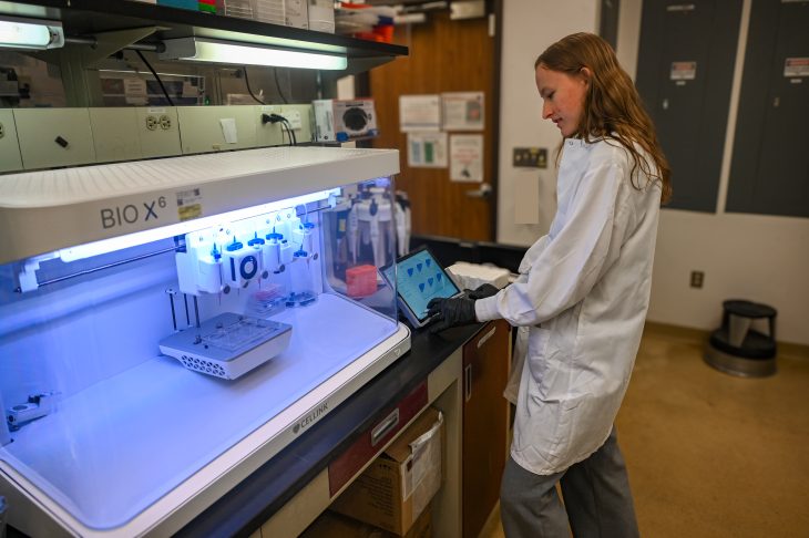 A student in a white lab coat and gloves operates a BIO X6 bioprinter using a tablet in a laboratory.