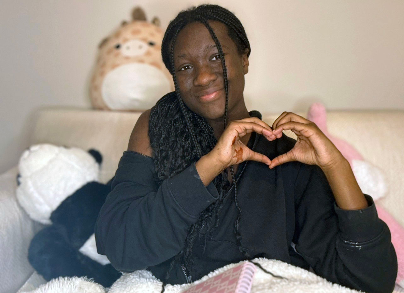 A girl with a black shirt and long black braids makes a heart cymbol with two hands. She is sitting on a bed with stuffed animals around her.