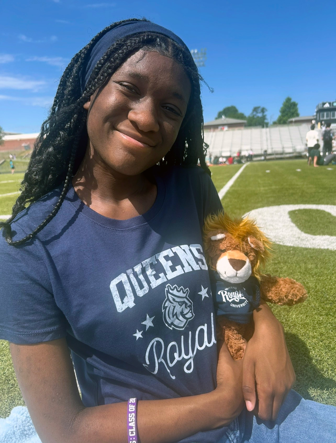 A girl with long brown braids stands on a football field with a navy blue Queens Royal teeshirt on holding a stuffed animal of Rex the Lion, the school mascot.