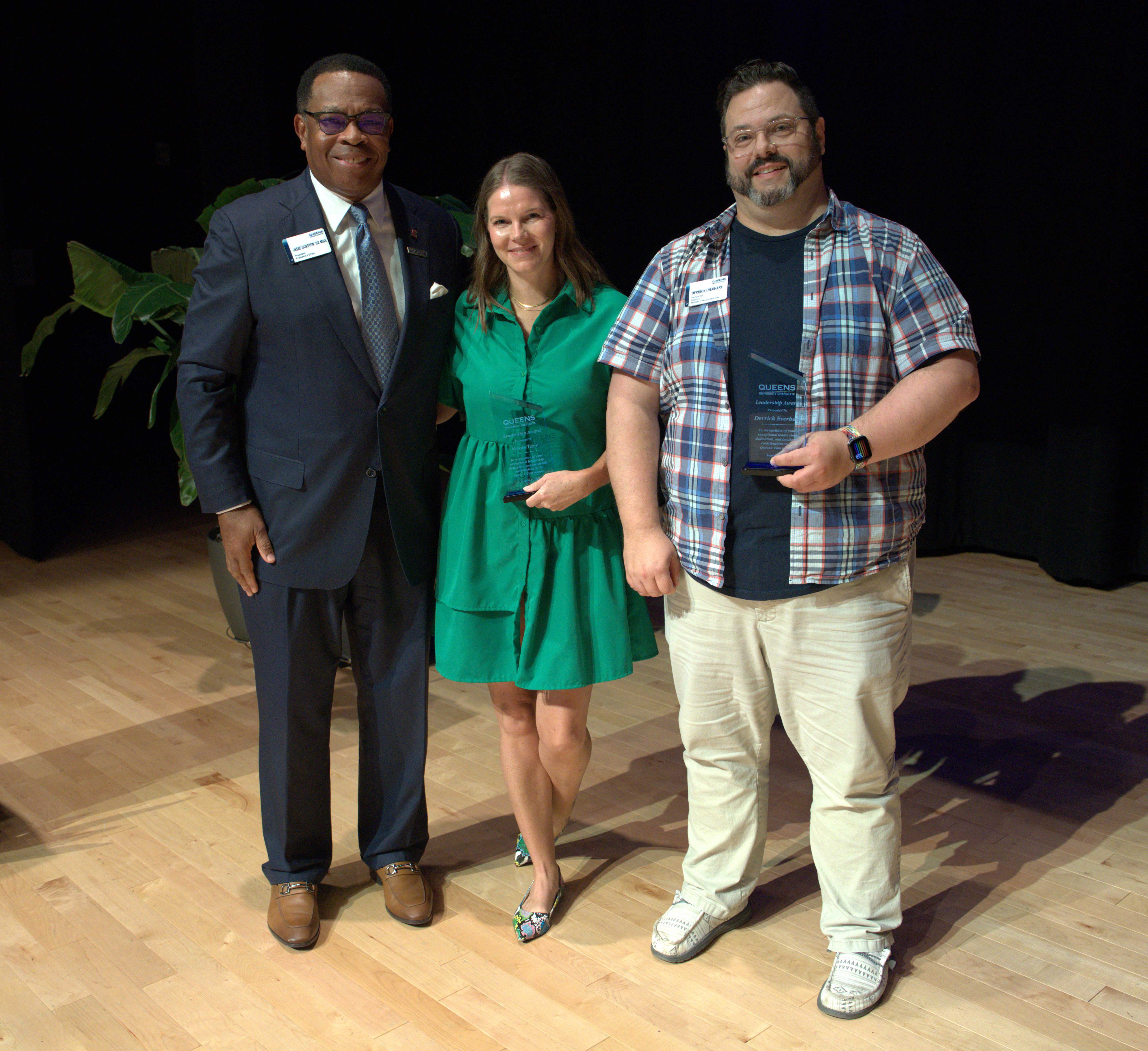 A photo of three people which includes a man with glasses in a dark suit stands on the left next to woman in a green dress holding a crystal award. Next to her on the right is a man with a plaid button down shirt and khaki pants holding a crystal award.