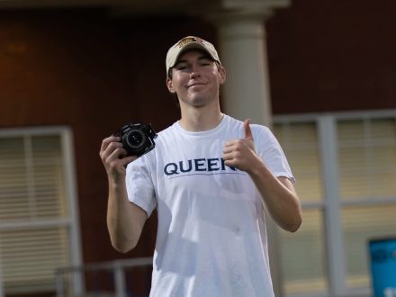 A smiling man with white baseball cap and teeshirt that says "Queens" holds a camera and gives a thumbs up.